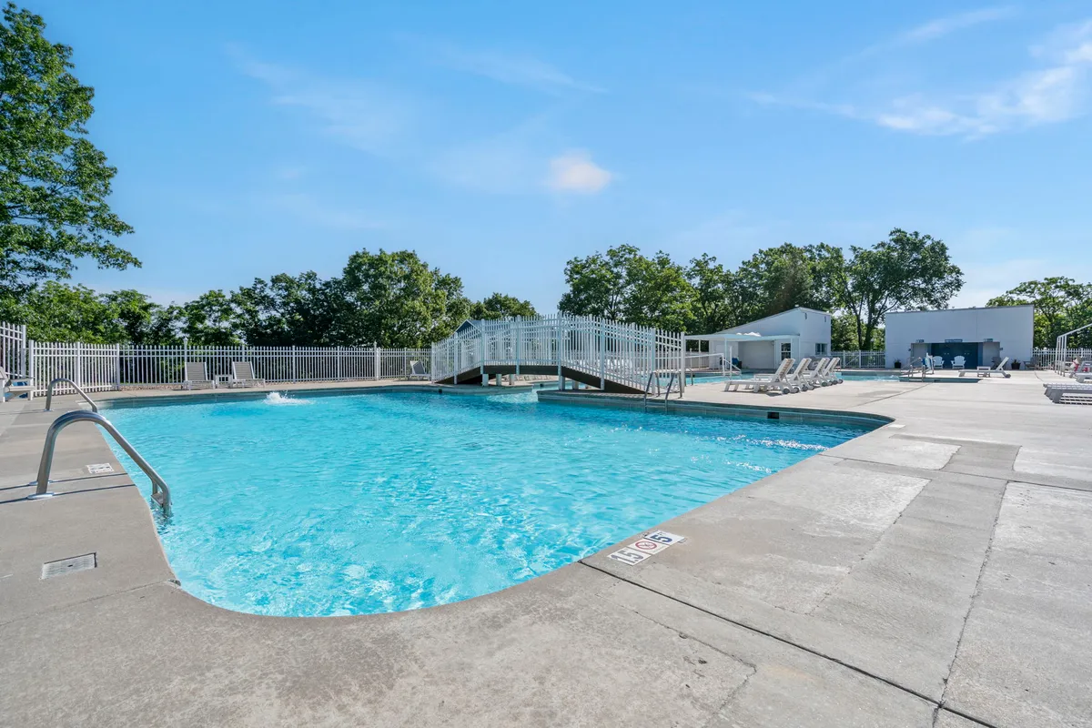A sparkling outdoor swimming pool bordered by a quaint bridge, comfortable lounge chairs, and well-kept greenery. The bright and cheerful mood invites guests to relax, swim, and socialize under the sun.
