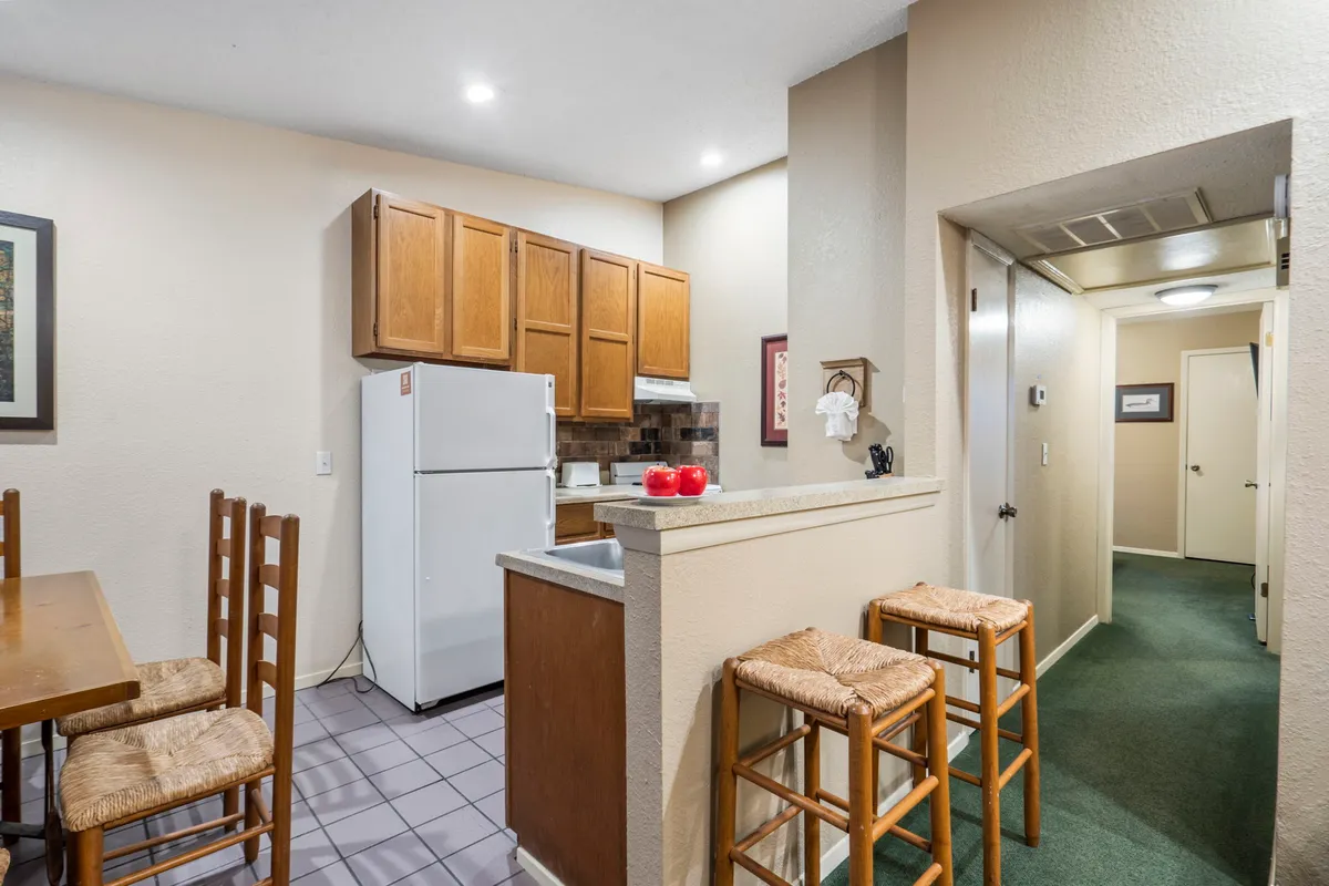 A welcoming kitchen and dining space featuring warm cabinetry, a dining table set for a meal, bar stools at the kitchen counter, and modern appliances including a refrigerator. The area is well-lit and designed for both cooking and gathering, with homey touches and plenty of space for meal prep and entertaining.