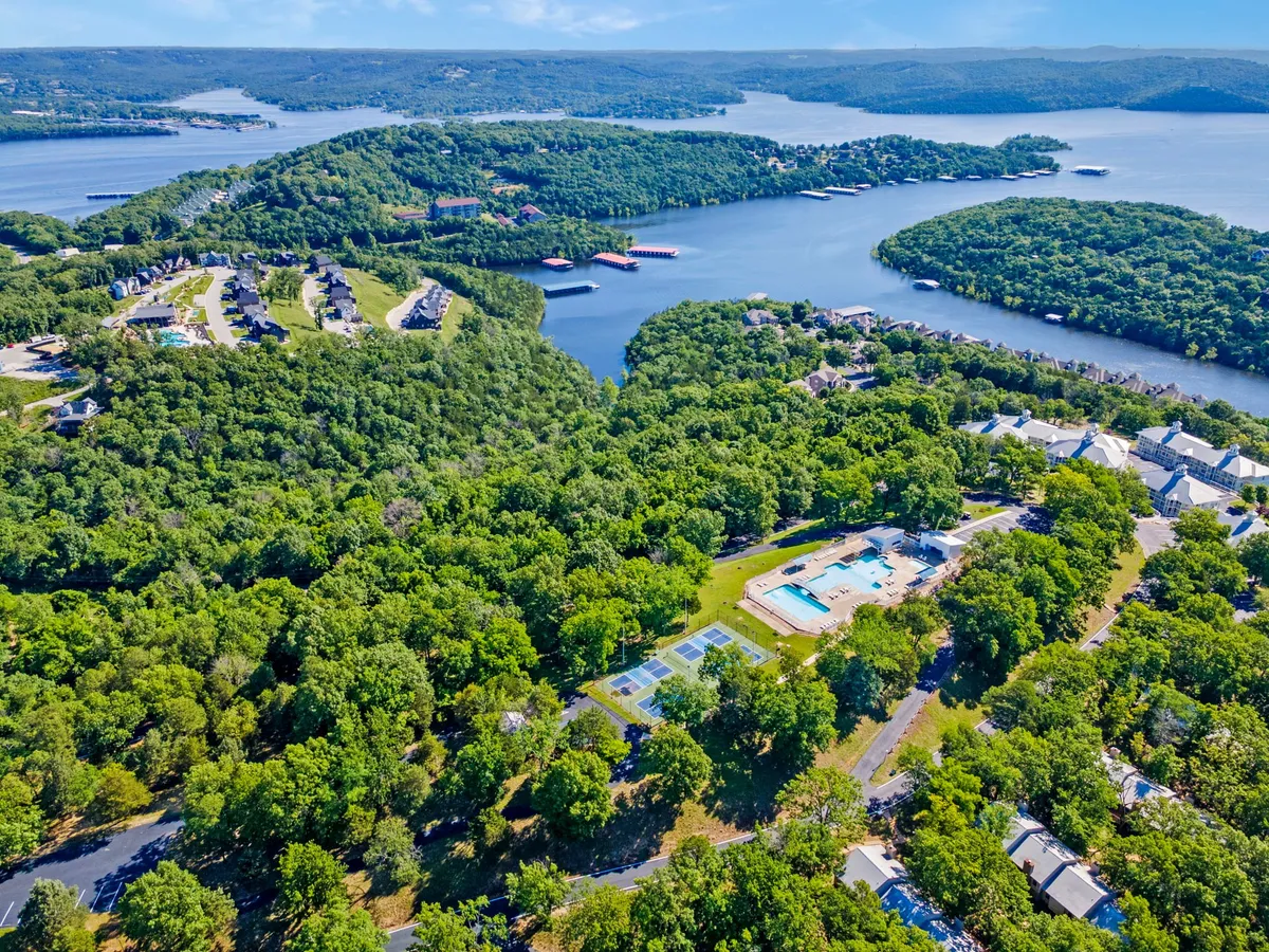 A sweeping view of the resort's amenities surrounded by lush greenery, including the lake, outdoor swimming pool, and tennis courts. The scene is vibrant and inviting, with clear skies enhancing the recreational vibe.