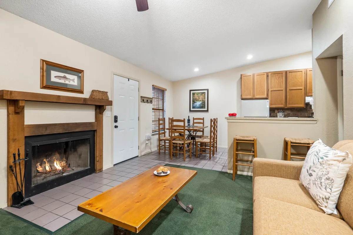 A wide-angle view of the living room, featuring a central fireplace, a comfortable couch, a dining table, and the adjacent kitchen area. Large windows let in natural light and offer a glimpse of the peaceful resort surroundings, while the open layout encourages social gathering.