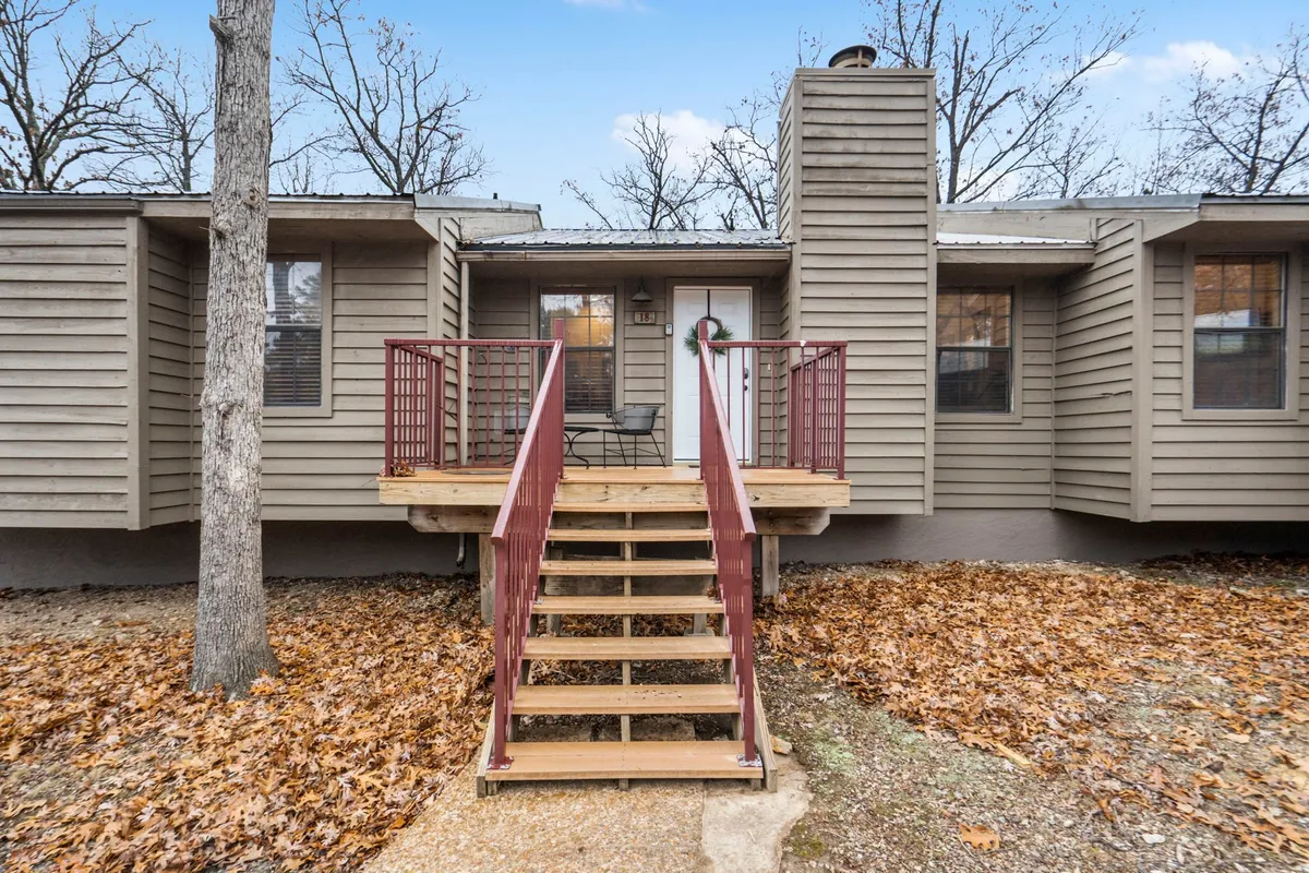 The exterior of a cozy residence is shown, featuring a welcoming deck area surrounded by trees, with a staircase leading up to the front door and a prominent chimney hinting at a fireplace inside. The setting feels secluded and peaceful—perfect for intimate outdoor relaxation or gatherings.