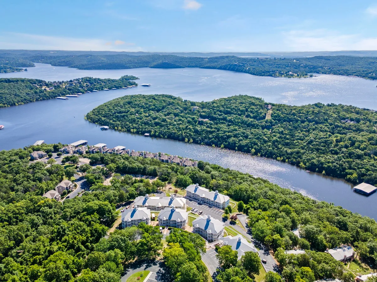 A tranquil, sweeping view of Table Rock Lake bordered by lush greenery, with residential buildings nestled along the shore and a winding river in the distance. The calm water invites boating and watersports, while the natural surroundings emphasize the lakefront setting.