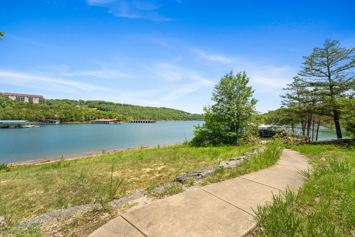 A scenic lakeside pathway edged with trees and gentle hills, leading toward the shimmering lake. People are seen walking and picnicking along the trail, with hints of fishing and boating activity in the distance. The tranquil setting is ideal for exploring nature with pets.