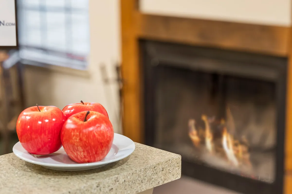 A cozy interior scene with three red apples on a plate placed on a table, with a glowing fireplace in the background. The warm setting invites guests to gather and unwind during cooler months.