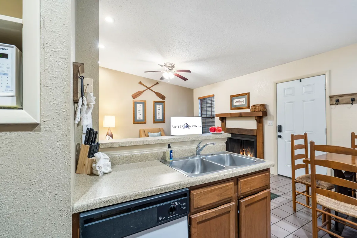 A view from the kitchen counter looking into the dining area and living room, with a fireplace as a focal point. Decorative oars and framed pictures add Ozark charm, while the layout emphasizes the seamless flow between kitchen, dining, and relaxation zones.