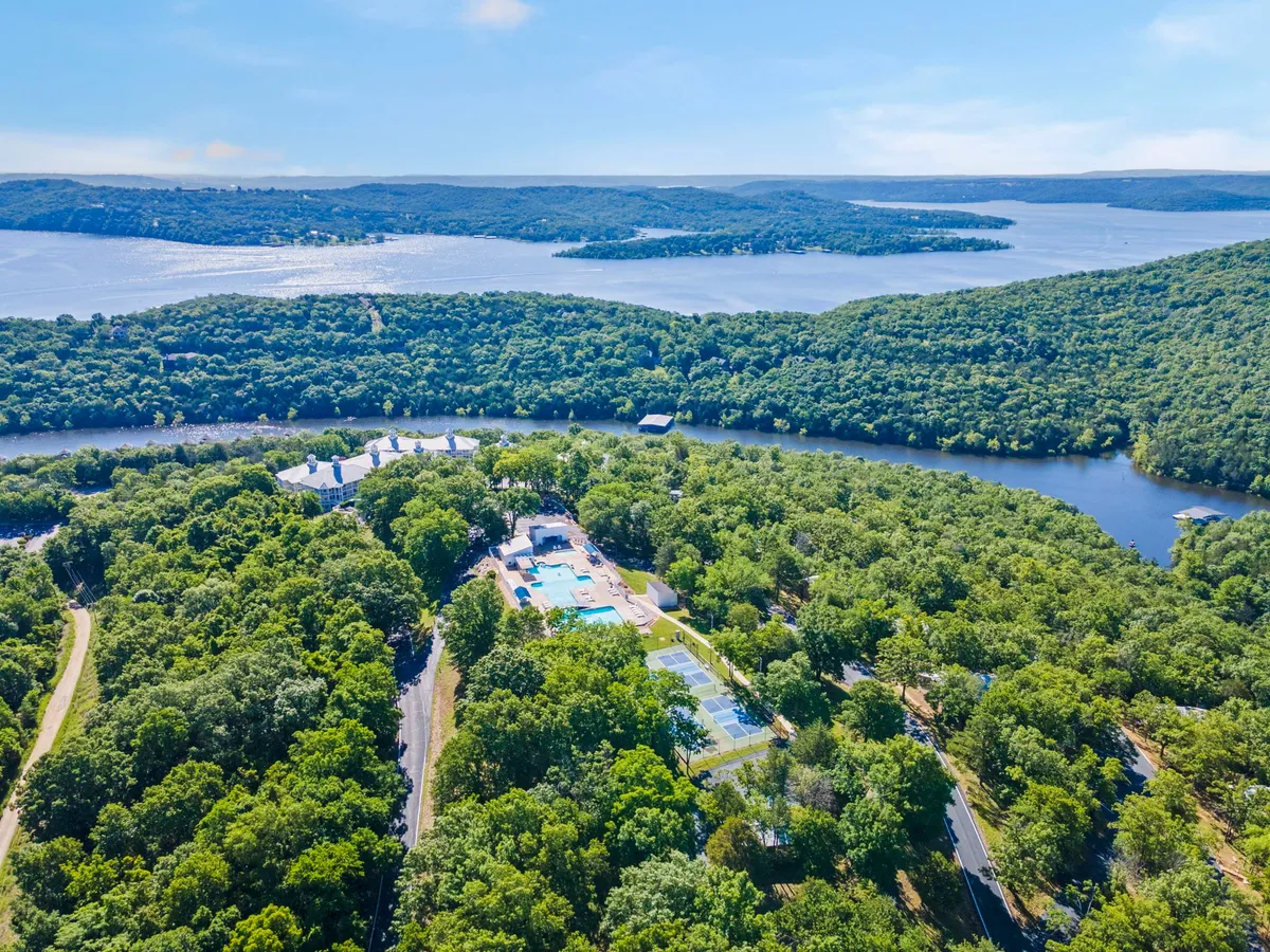 This image captures an expansive view of the lake framed by lush green trees, with a swimming pool area and tennis courts visible in the foreground. The atmosphere is serene and inviting, offering a panoramic perspective of the natural surroundings and the amenities available for exploration and relaxation.