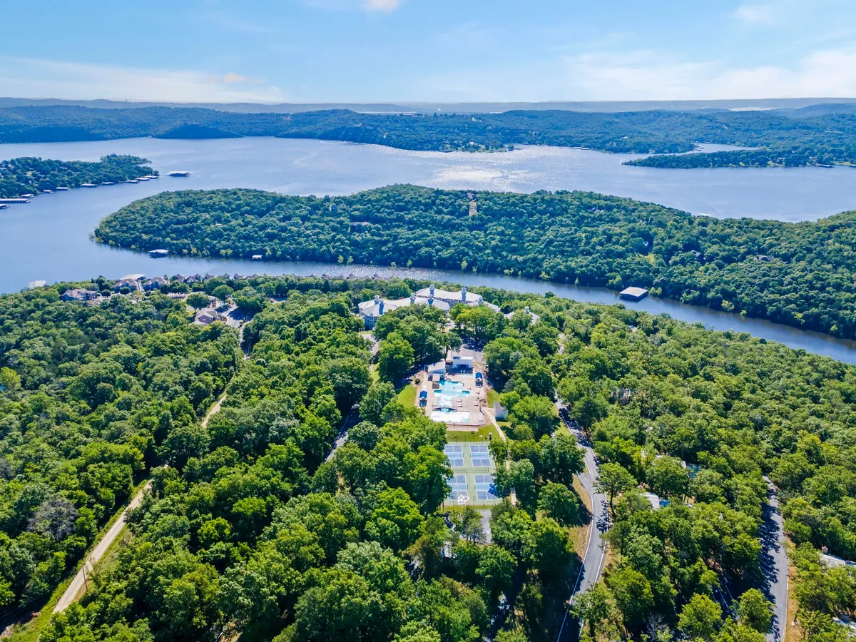 A vibrant exterior shot capturing lush greenery, a serene lake view, a sparkling swimming pool, and tennis courts. The scenery is bathed in natural light, with hints of recreational activities possible in every corner.