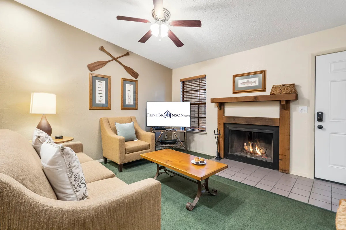 The image showcases a cozy interior living space with a warm fireplace as the focal point, surrounded by comfortable seating, a television for entertainment, decorative paddles that hint at the nearby lake, and a wooden coffee table perfect for games or gathering. The inviting setup is ideal for relaxing evenings with family or friends.