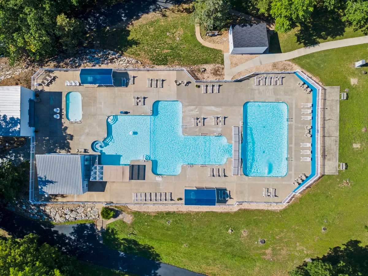 A lively exterior scene of a sparkling swimming pool bordered by deck chairs and surrounded by lush greenery. The sun is beginning to set, casting a warm glow over the pool area, suggesting an inviting atmosphere for swimming, sunbathing, and socializing during warm weather.