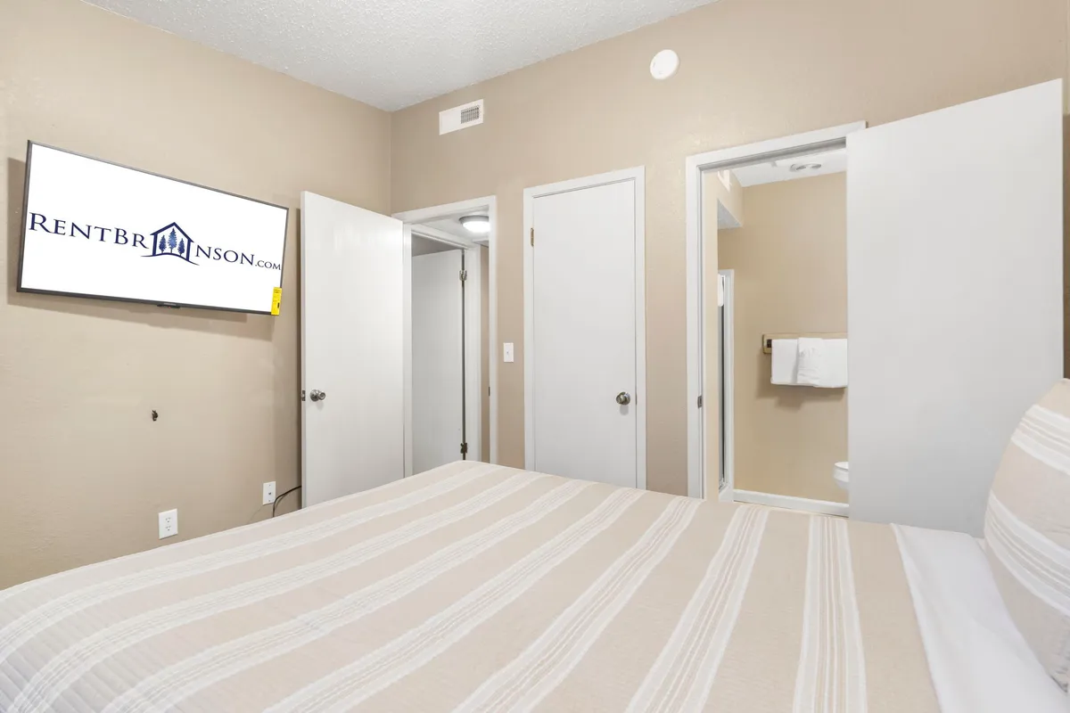 A bedroom with a bed dressed in striped linens, a wall-mounted television, and a visible doorway leading to an en suite bathroom. The scene is inviting and highlights the convenience of having private bathroom access.