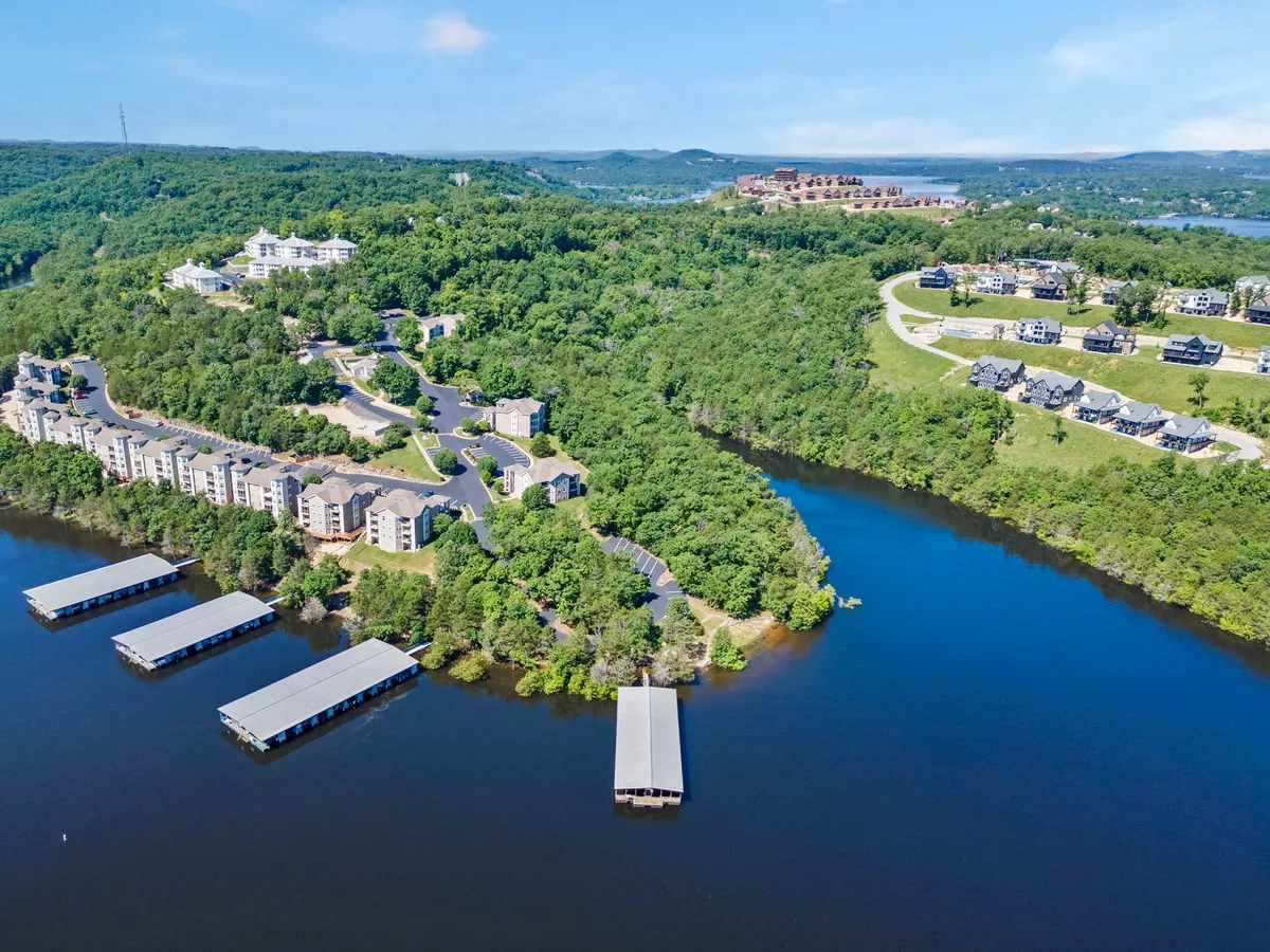 A peaceful view of a lakefront area dotted with docks, residential buildings, and lush greenery. People are seen relaxing by the water, engaging in boating and fishing, with hills framing the tranquil scene. The ambiance conveys a resort-style lake experience perfect for every season.